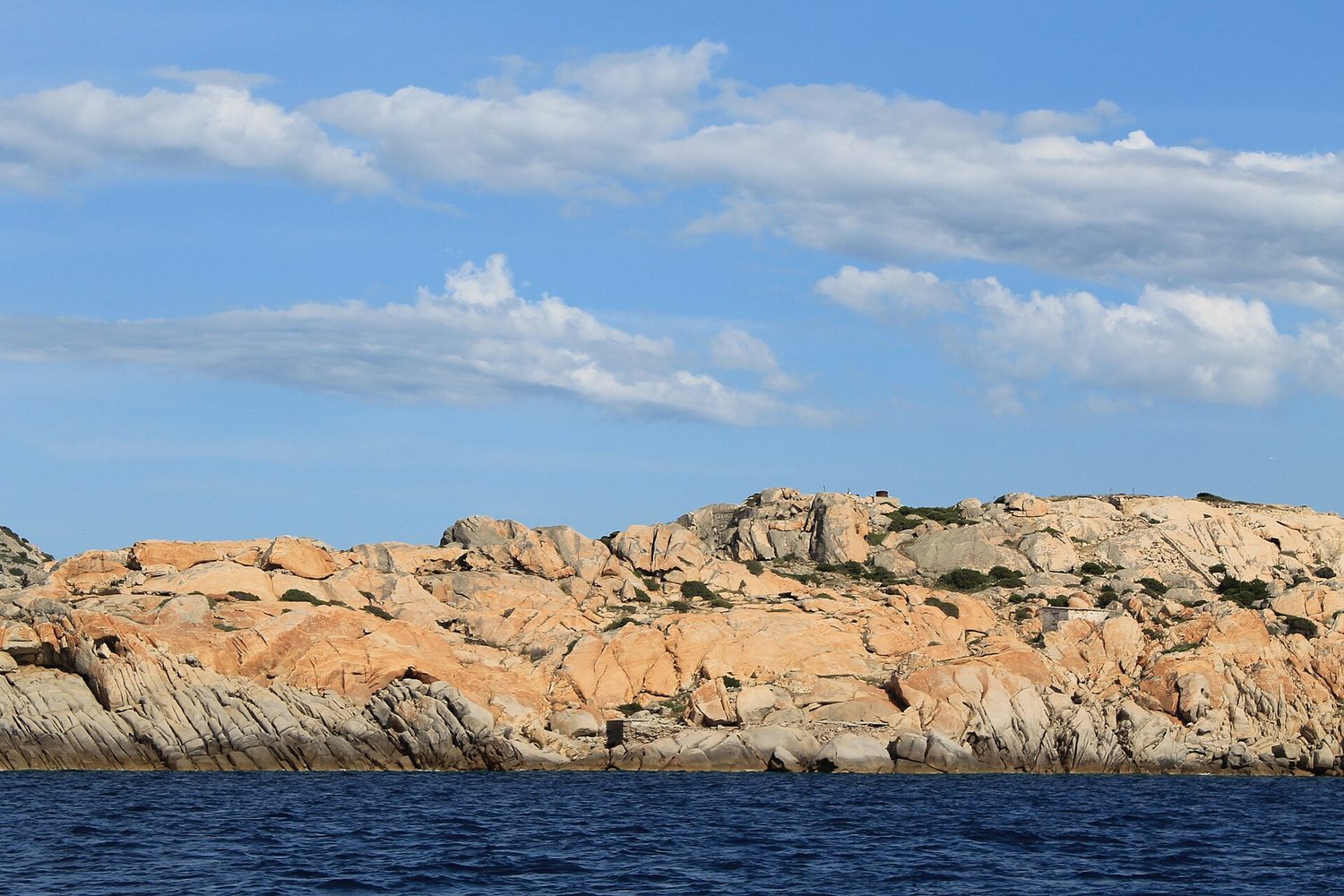 Costa di un'isola dell'Arcipelago di La Maddalena vista dal mare: rocce di granito rosato levigate dal vento, bassa macchia mediterranea e acqua blu scuro